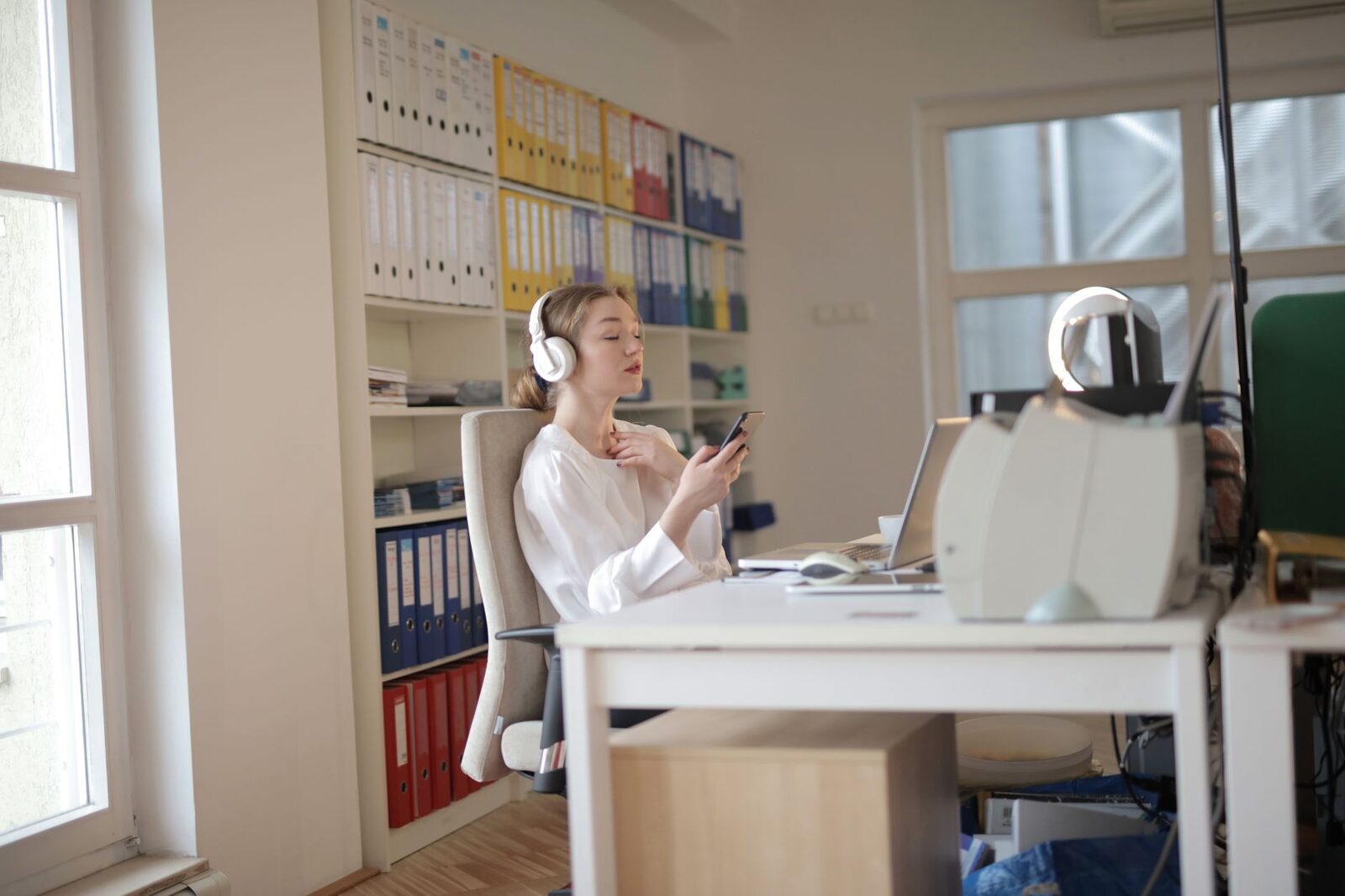 Woman sitting at desk in modern office, using smartphone and headphones.